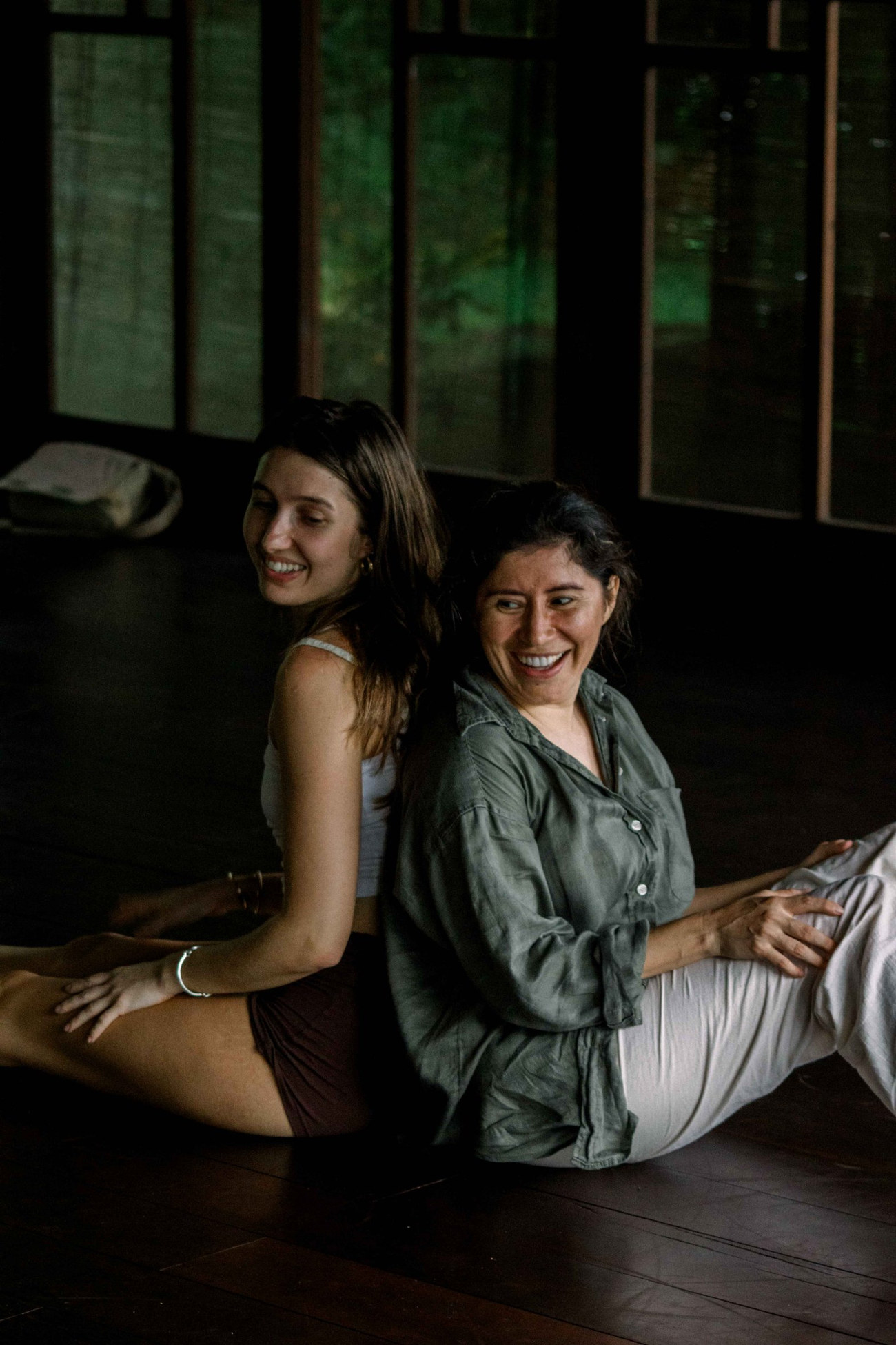 Two dancers sitting back to back on a dark wooden floor, laughing